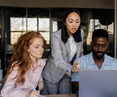 Three people collaboratively work at a laptop. One man is coding, while two are making suggestions.