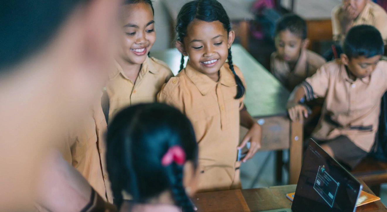 Children stand around a laptop smiling, while one child works with her back to us on the laptop.